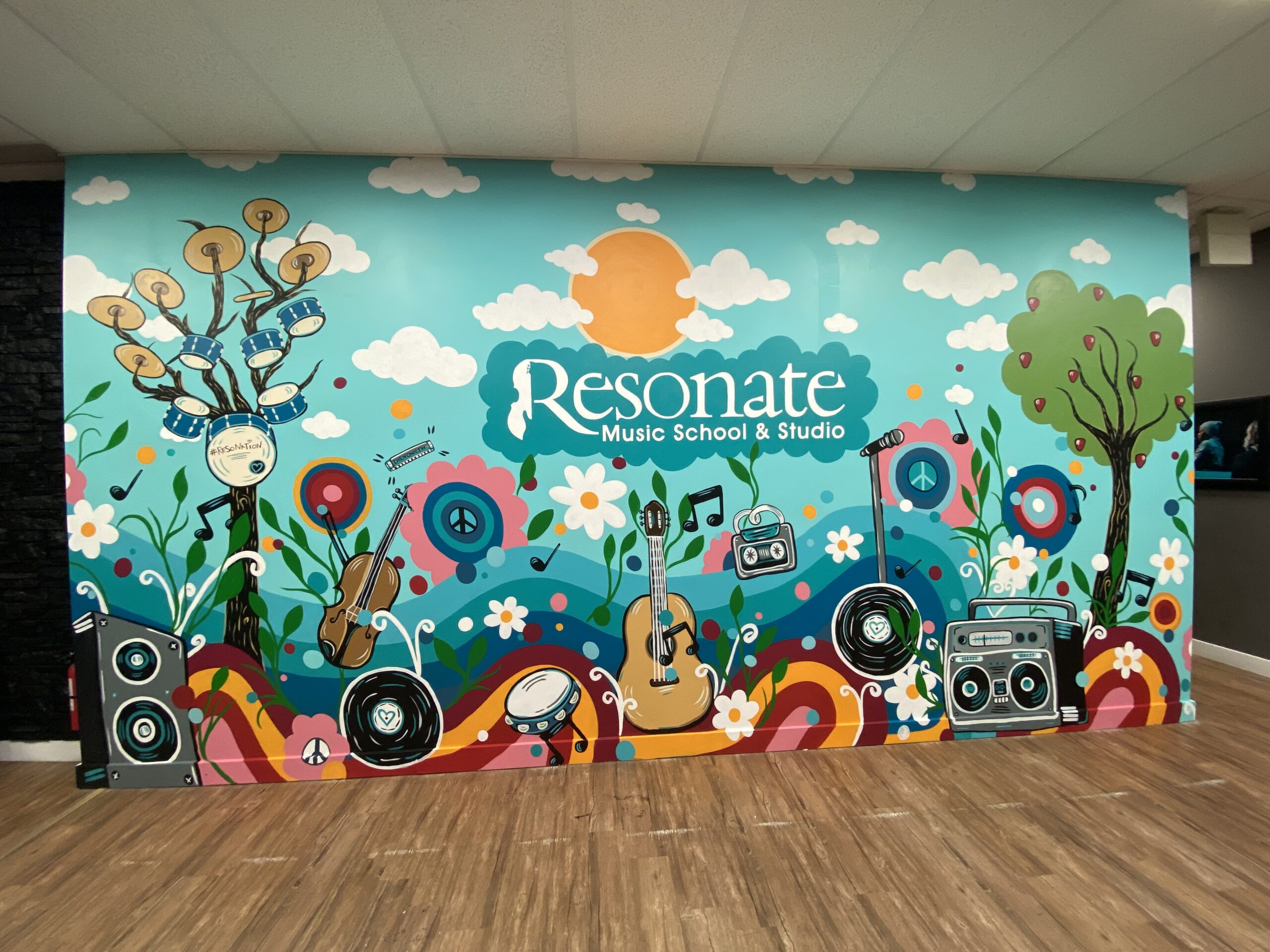 Colorful and expressive mural inspired by music. Hand painted mural by Carly Marie Artistry at Resonate Music School & Studio in north Edmonton. Mural features instruments and trees and flowers with bright brand color teal and clouds with a sun and the name of the school in white