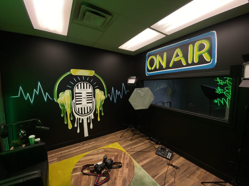 Podcast Room Just Bite Me black walls with custom painted mural artwork by Edmonton Artist Carly Marie. Bright colors handpainted microphone with headphones and sound lines coming out of it
