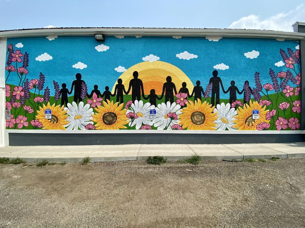 Large scaled community inspired mural hand painted by Edmonton artist Carly Marie. Mural is painted on Onoway Community Hall facing the parking lot featuring black silhouettes of people holding hands, large colorful flowers, blue skies with white clouds